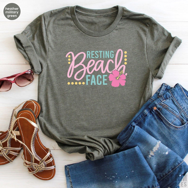 May include: A heather military green t-shirt with the text "Resting Beach Face" in pink, blue, and yellow lettering. A pink hibiscus flower is to the right of the text.
