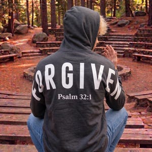 May include: A dark gray hoodie with the word "FORGIVE" printed in large white letters across the back. Below the word is the text "Psalm 32:1". The person is wearing blue jeans and is sitting outdoors.