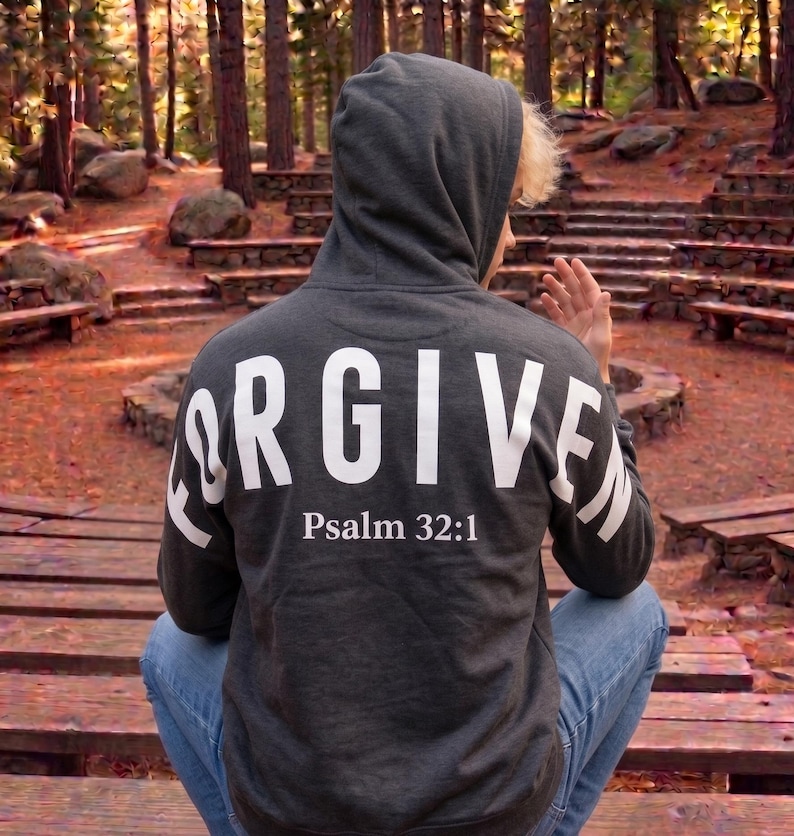 May include: A dark gray hoodie with the word "FORGIVE" printed in large white letters across the back. Below the word is the text "Psalm 32:1". The person is wearing blue jeans and is sitting outdoors.