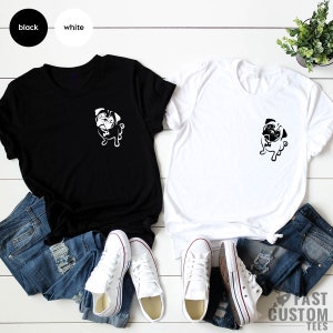 May include: Two t-shirts featuring a black and white pug graphic on the front. The t-shirts are laid out on a white wooden surface with a pair of jeans and white trainers on each side.