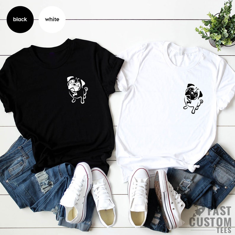 May include: Two t-shirts featuring a black and white pug graphic on the front. The t-shirts are laid out on a white wooden surface with a pair of jeans and white trainers on each side.