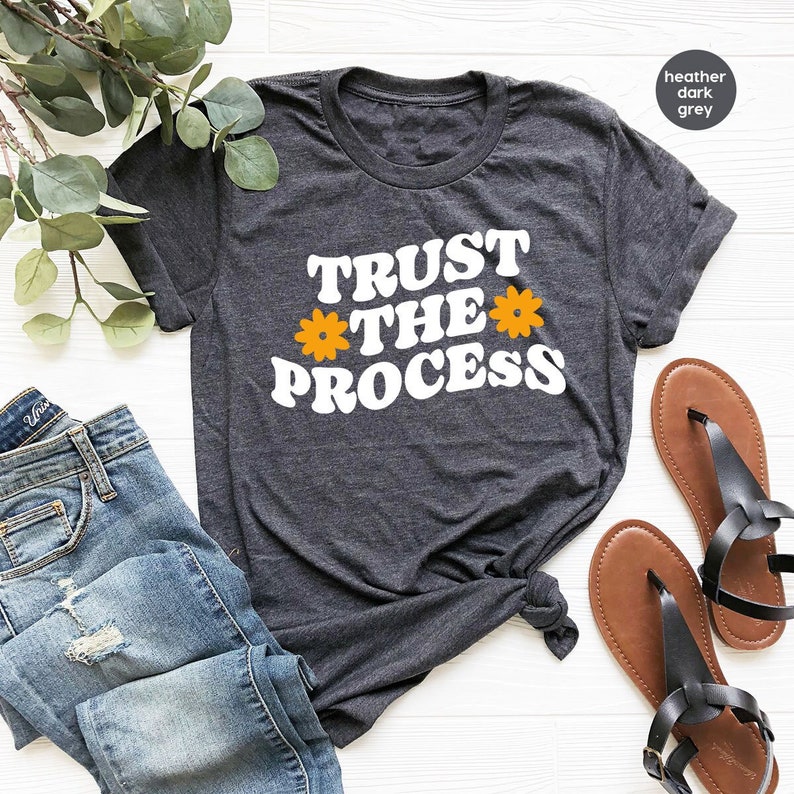 May include: A heather dark grey t-shirt with the text "TRUST THE PROCESS" in white with two yellow flower graphics.