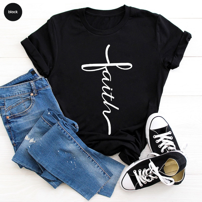 May include: Black t-shirt with a white cross and the word "faith" written vertically on it. The shirt is laid out on a wooden surface with a pair of blue jeans and black sneakers.