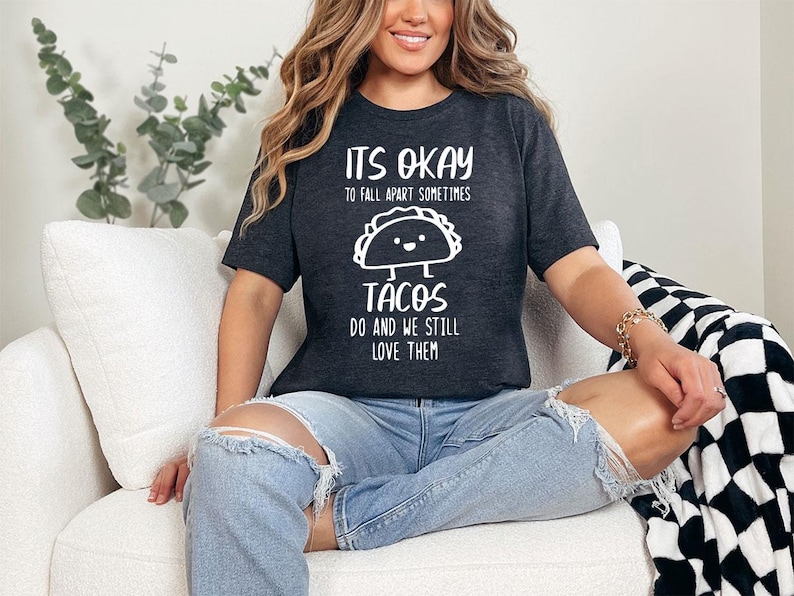 May include: A woman wearing a dark grey t-shirt with the text "It's okay to fall apart sometimes Tacos do and we still love them" and a cartoon taco graphic. She is sitting on a white couch with a black and white checkered throw blanket.