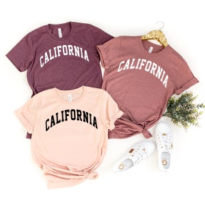 May include: Three t-shirts with the word "CALIFORNIA" printed in white on a maroon shirt and a peach shirt, and in black on a light pink shirt.