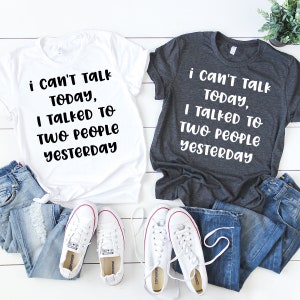 May include: Two white t-shirts with black text that reads "i can't talk today, I talked to two people yesterday". One shirt is white and the other is dark grey. The shirts are laid out on a white surface with a pair of white sneakers and two pairs of blue jeans.