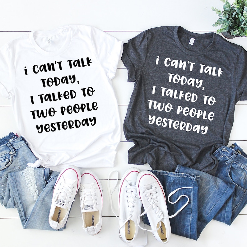 May include: Two white t-shirts with black text that reads "i can't talk today, I talked to two people yesterday". One shirt is white and the other is dark grey. The shirts are laid out on a white surface with a pair of white sneakers and two pairs of blue jeans.