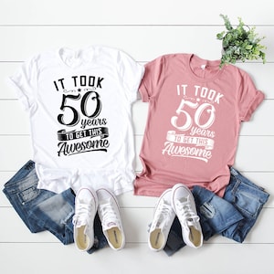 May include: Two white t-shirts with black text that reads "It took 50 years to get this Awesome". One shirt is on a pink background and the other is on a white background. Both shirts are paired with blue jeans and white sneakers.