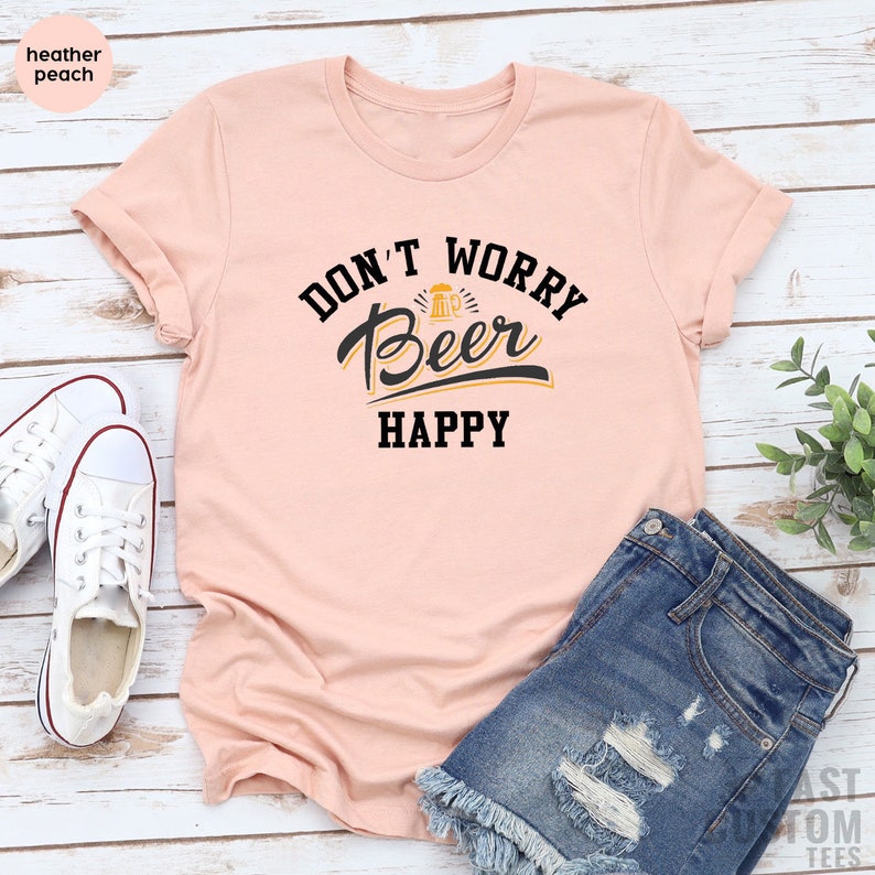 May include: A peach colored t-shirt with the text "Don't Worry Beer Happy" in black lettering. The text includes a stylized beer mug graphic.