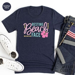 May include: A heather navy blue t-shirt with the text "Resting Beach Face" in pink and teal lettering with a pink hibiscus flower graphic.