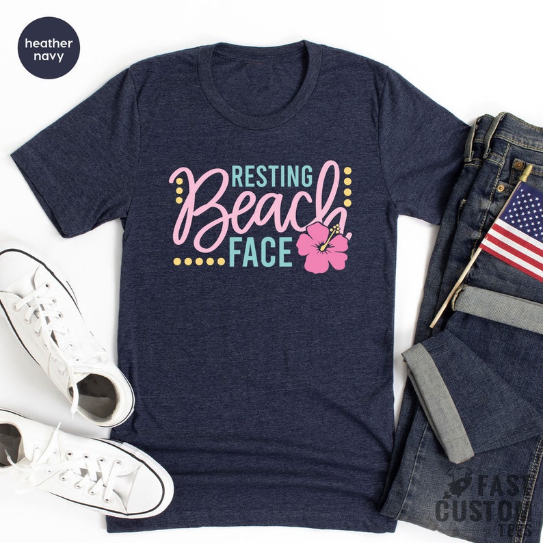 May include: A heather navy blue t-shirt with the text "Resting Beach Face" in pink and teal lettering with a pink hibiscus flower graphic.