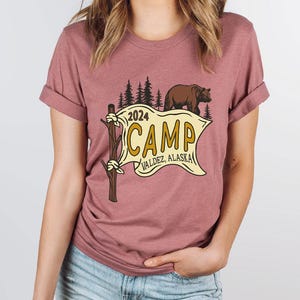 a woman wearing a t - shirt that says camp with a bear and a banner