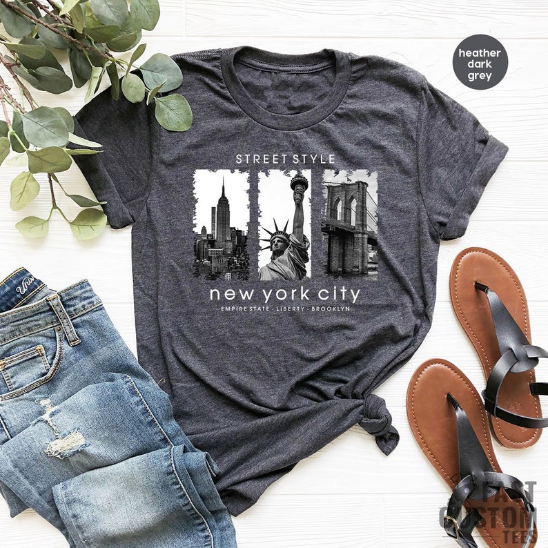 May include: Heather dark grey t-shirt with a graphic print of the New York City skyline, including the Empire State Building, the Statue of Liberty, and the Brooklyn Bridge. The text "Street Style" and "New York City" is printed on the shirt. The text "Empire State, Liberty, Brooklyn" is printed below the graphic.