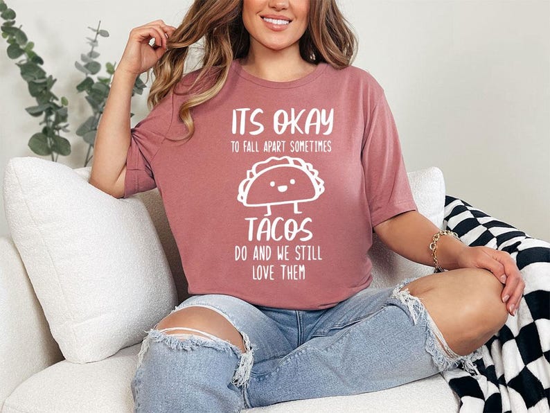 May include: A woman wearing a pink t-shirt with the text "It's okay to fall apart sometimes tacos do and we still love them" and a cartoon taco graphic. She is sitting on a white couch with a black and white checkered blanket.
