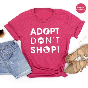 May include: Heather raspberry t-shirt with the text "Adopt Don't Shop!" in white. The text is surrounded by circles with a cat and dog silhouette inside each circle.