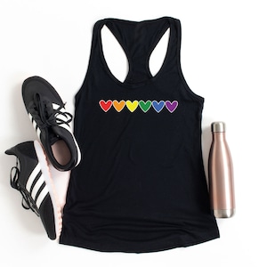 May include: Black tank top with a rainbow of hearts across the front. The tank top is lying flat on a white surface with a pair of black and white trainers and a rose gold water bottle.