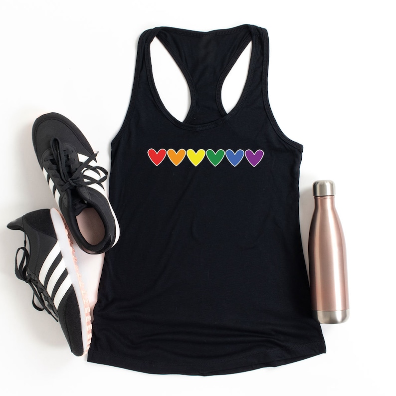 May include: Black tank top with a rainbow of hearts across the front. The tank top is lying flat on a white surface with a pair of black and white trainers and a rose gold water bottle.