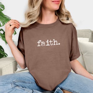 May include: A brown short-sleeved t-shirt featuring the word faith. printed in a white, distressed, typewriter-style font across the centre of the chest. The garment has a crew neckline and a relaxed fit, constructed from a solid-coloured fabric.