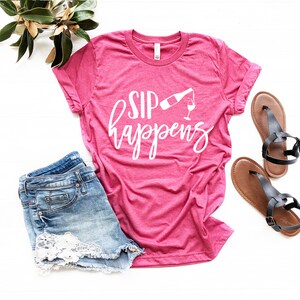 May include: Pink t-shirt with white text that says "Sip Happens" and a graphic of a wine bottle pouring into a wine glass.