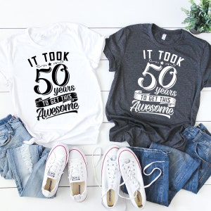 May include: Two t-shirts, one white and one dark gray, with the text "It took 50 years to get this awesome" printed on them. The shirts are laid out on a white wooden surface with a pair of white sneakers and two pairs of blue jeans.