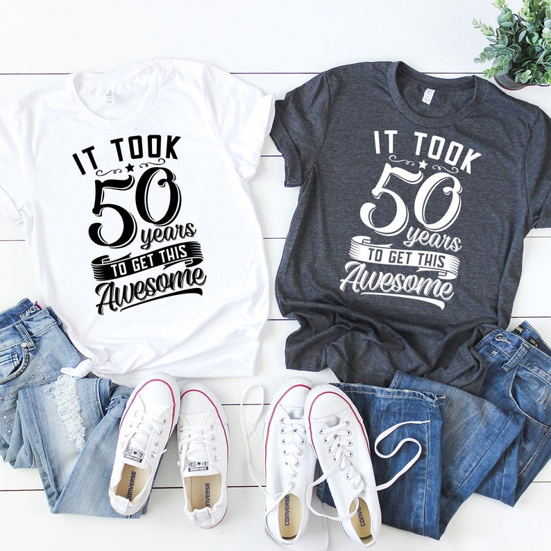 May include: Two t-shirts, one white and one dark gray, with the text "It took 50 years to get this awesome" printed on them. The shirts are laid out on a white wooden surface with a pair of white sneakers and two pairs of blue jeans.
