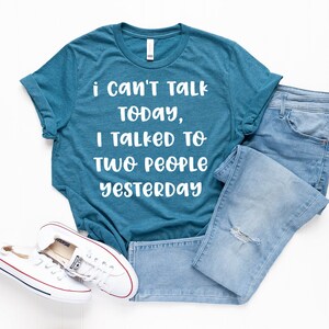 May include: A teal blue t-shirt with white text that reads "i can't talk today, i talked to two people yesterday". The shirt is laid flat on a white surface with a pair of light blue jeans and white sneakers.