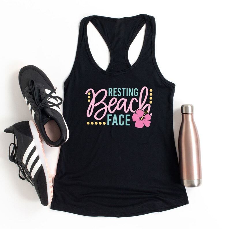 May include: Black tank top with the text "Resting Beach Face" in a colorful font with a pink hibiscus flower. The tank top is laying on a white surface with a pair of black and white sneakers and a rose gold water bottle.