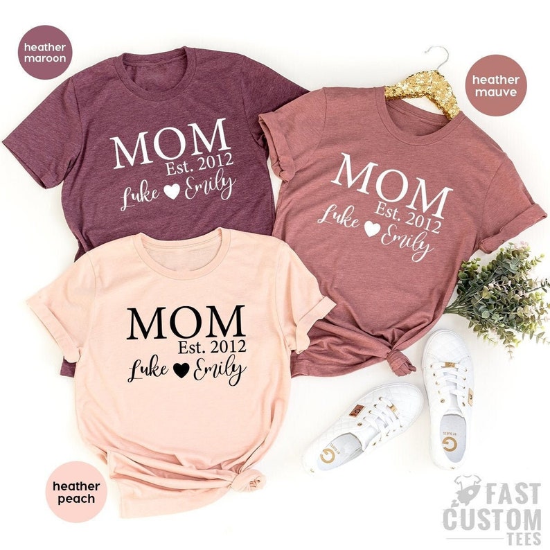 May include: Three t-shirts in different colours with the text "MOM Est. 2012 Luke Emily" on them. The shirts are heather maroon, heather peach, and heather mauve.