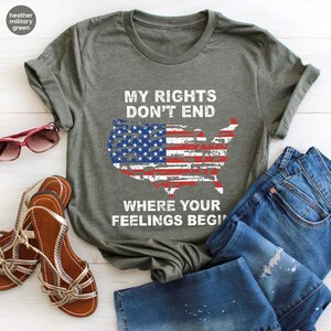 May include: Heather military green t-shirt with the text "MY RIGHTS DON'T END WHERE YOUR FEELINGS BEGIN" and an American flag design in the shape of the United States. The shirt is paired with blue jeans, sandals, and sunglasses.