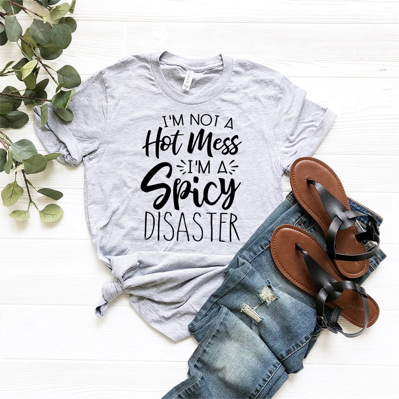 May include: A gray t-shirt with black text that reads "I'm not a hot mess I'm a spicy disaster". The shirt is laid out on a white surface with a pair of blue jeans and brown sandals.