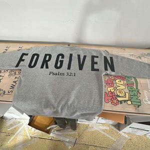 May include: A gray sweatshirt with the word "FORGIVEN" printed in large black letters across the chest, with "Psalm 32:1" below. The long-sleeved shirt is laid out on a table with other printed materials.