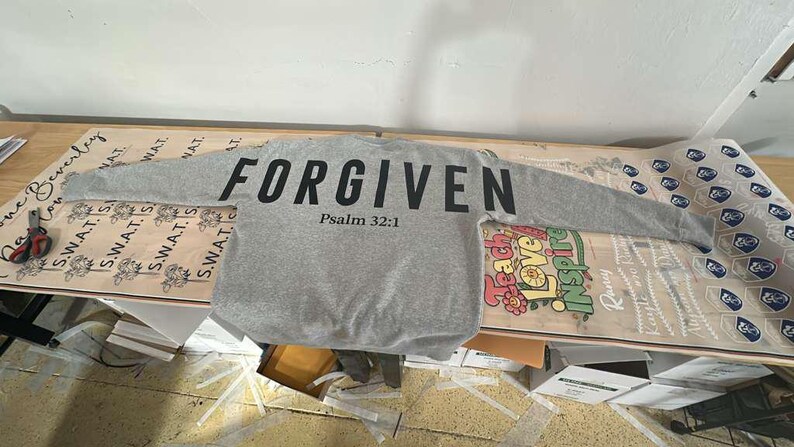 May include: A gray sweatshirt with the word "FORGIVEN" printed in large black letters across the chest, with "Psalm 32:1" below. The long-sleeved shirt is laid out on a table with other printed materials.