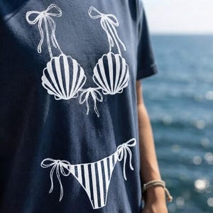 Peut inclure: Un t-shirt bleu marine avec une impression graphique blanche représentant un bikini. La partie supérieure affiche un soutien-gorge en forme de coquillage avec des liens à nouer, et le bas montre une culotte à rayures verticales.