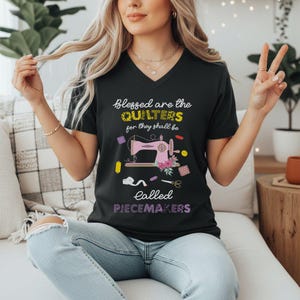 May include: Black V-neck t-shirt with the text "Blessed are the QUILTERS for they shall be called PIECEMAKERS." The shirt features a pink sewing machine graphic with floral accents and sewing notions.