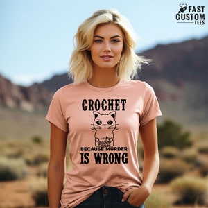 Op de afbeelding: Een roze T-shirt met een zwarte katillustratie en de tekst "Haakwerk omdat moord verkeerd is".