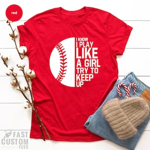 Op de afbeelding: Rood T-shirt met een witte baseball-afbeelding en de tekst "I know I play like a girl try to keep up".
