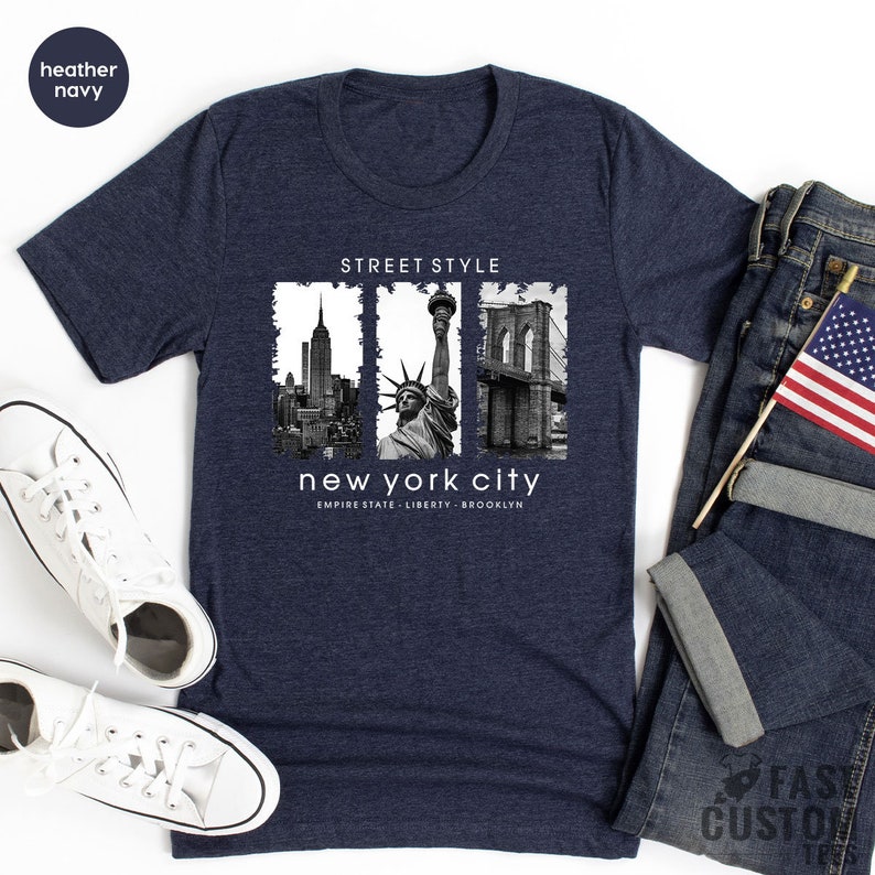 May include: A heather navy blue t-shirt with a graphic print of the New York City skyline, the Statue of Liberty, and the Brooklyn Bridge. The text "Street Style" and "new york city" is printed on the shirt. The text "Empire State - Liberty - Brooklyn" is printed below the graphic.