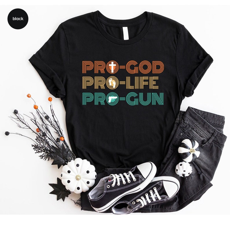 May include: Black t-shirt with the text "PRO-GOD PRO-LIFE PRO-GUN" in orange, yellow, and teal. The shirt features a cross, a fetus, and a handgun graphic. The shirt is paired with black jeans and black trainers.