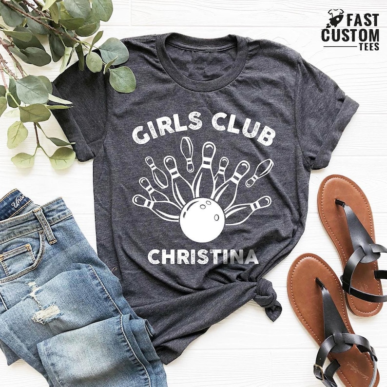 Pode incluir: Uma camiseta cinza escura com o texto "Girls Club" e um gr&aacute;fico de uma bola de boliche e pinos. O nome "Christina" est&aacute; impresso abaixo do gr&aacute;fico.