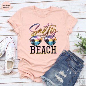 May include: A pink t-shirt with the text "Salty Beach" in a colorful, glittery font. The text is overlaid with a pair of sunglasses with a beach scene reflected in the lenses.
