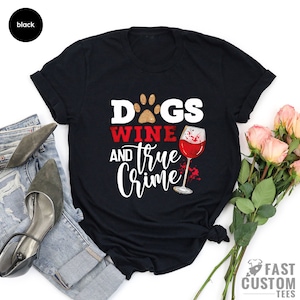 May include: Black t-shirt with the words "DOGS, WINE, and TRUE CRIME" in white and red text. A paw print and a wine glass graphic are also featured. The shirt is a casual wear item.