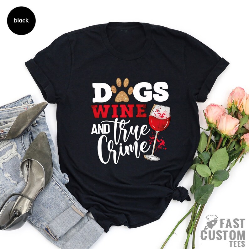 May include: Black t-shirt with the words "DOGS, WINE, and TRUE CRIME" in white and red text. A paw print and a wine glass graphic are also featured. The shirt is a casual wear item.