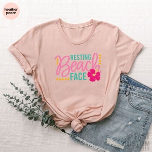 May include: A heather peach t-shirt with the text "Resting Beach Face" in pink, teal, and yellow lettering with a pink hibiscus flower graphic.