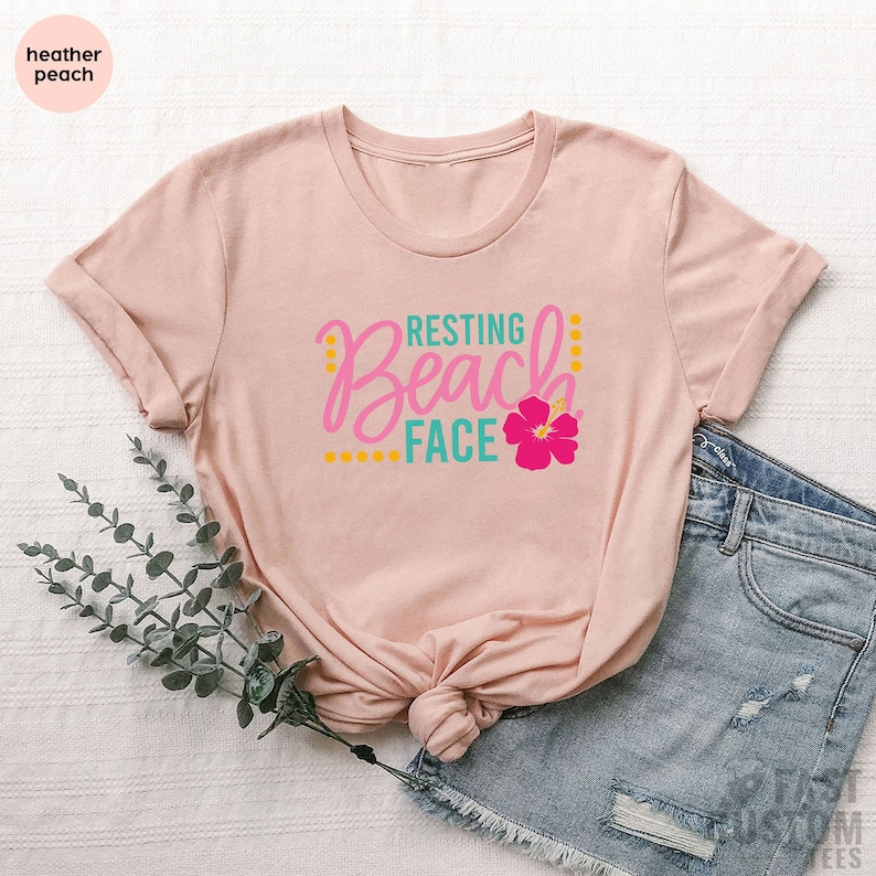 May include: A heather peach t-shirt with the text "Resting Beach Face" in pink, teal, and yellow lettering with a pink hibiscus flower graphic.