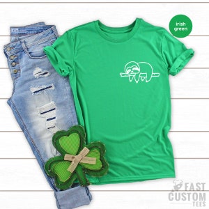 K&ouml;nnte beinhalten: Ein gr&uuml;nes T-Shirt mit einem wei&szlig;en Faultier-Motiv auf der Vorderseite. Das Shirt liegt auf einer wei&szlig;en Holzoberfl&auml;che mit einer blauen Jeans und einem gr&uuml;nen Kleeblatt.