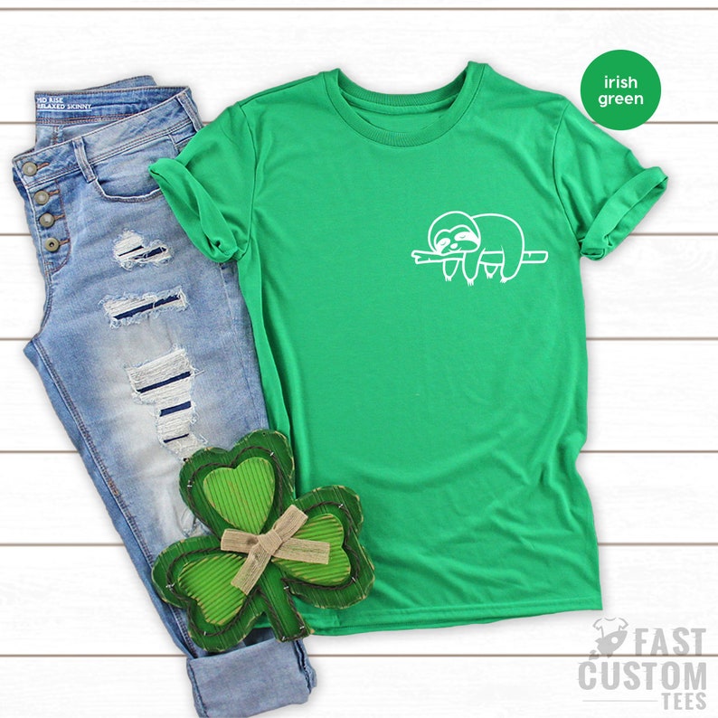 K&ouml;nnte beinhalten: Ein gr&uuml;nes T-Shirt mit einem wei&szlig;en Faultier-Motiv auf der Vorderseite. Das Shirt liegt auf einer wei&szlig;en Holzoberfl&auml;che mit einer blauen Jeans und einem gr&uuml;nen Kleeblatt.