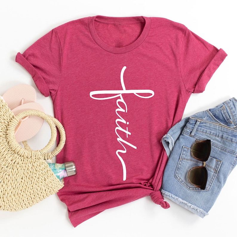 May include: A red t-shirt with a white cross and the word "faith" written vertically on it. The shirt is laid out on a white surface with a straw bag, flip flops, and denim shorts.