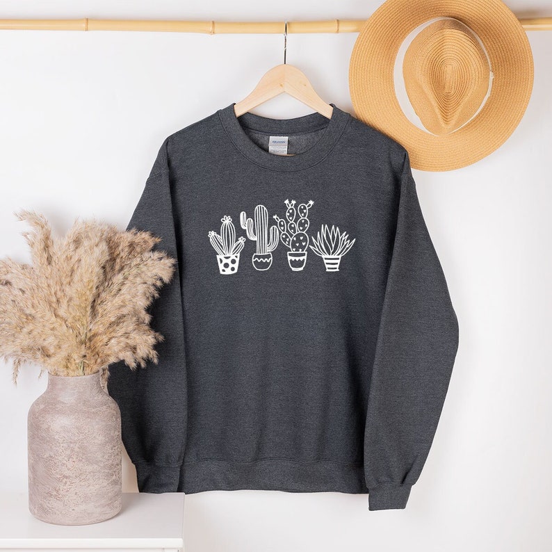 May include: Dark gray sweatshirt featuring a white graphic of four potted cacti. The cacti are in different shapes and sizes, with detailed white outlines. The sweatshirt is hanging on a wooden hanger against a neutral background, with a straw hat visible.