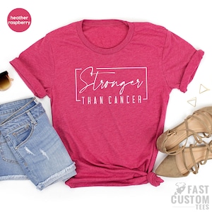 May include: A heather raspberry t-shirt featuring the white text "Stronger Than Cancer" in a stylish font. The sleeves are rolled up, and the bottom is knotted. The shirt is styled with denim shorts and sandals.
