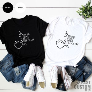 Pode incluir: Duas camisetas, uma preta e uma branca, com a palavra "She" escrita em letra cursiva e as palavras "Strong, Fierce, Brave, Full of Fire" empilhadas verticalmente abaixo.